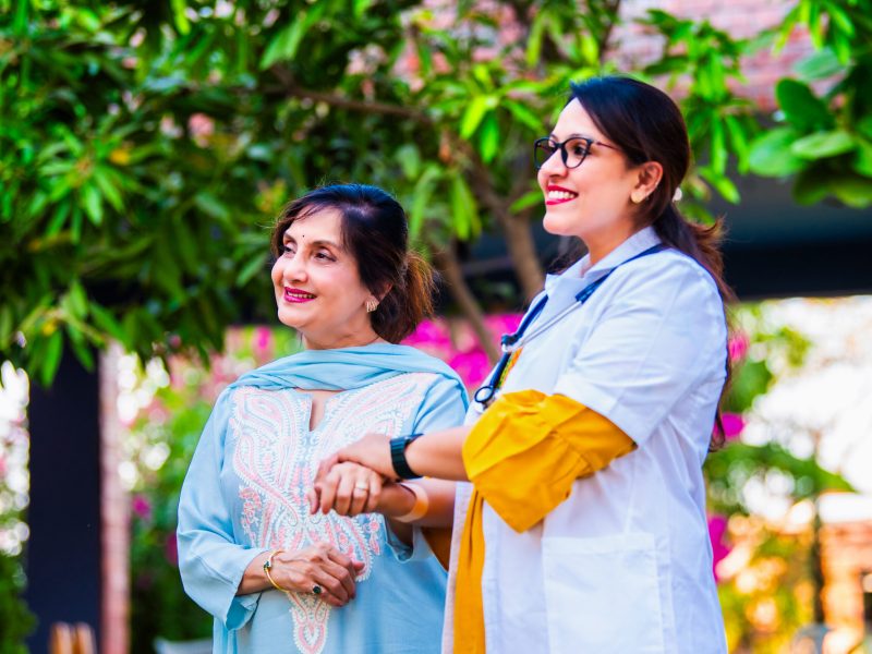 Indian Asian young female doctor standing with a senior woman patient outdoors in hospital premises, both smiling at the camera, conveying trust and care, with trees and buildings in the background